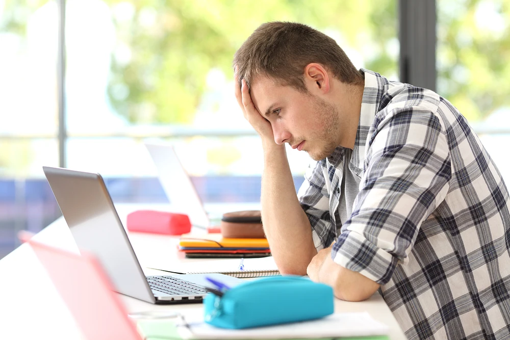 A student sits at a table looking frustrated at his laptop